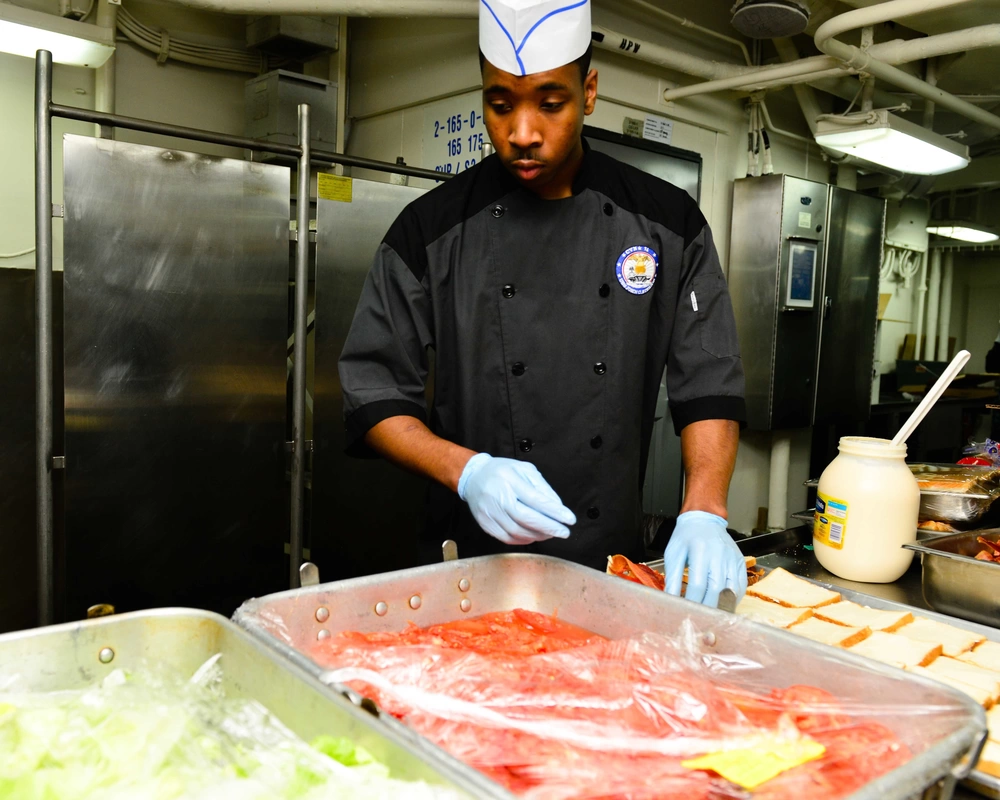 a Navy soldier makes sandwiches with bread, mayonnaise, lettuce and tomatoes.