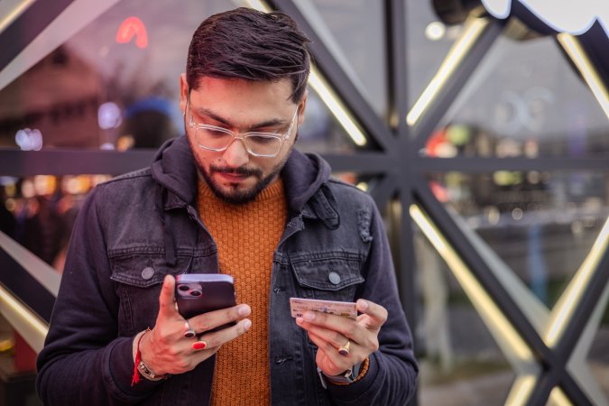 During his winter break, a young man from India wearing casual clothing and glasses can be seen shopping online on a busy street with a glass building in the background.